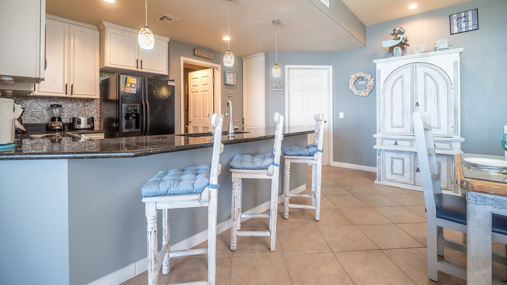 A kitchen with white cabinets, a black refrigerator, a coffee maker, and a sink. There are three white chairs with blue cushions.