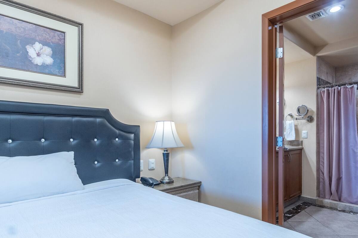 A hotel room with a bed, a bedside lamp, a framed picture on the wall, and a bathroom visible through an open door.