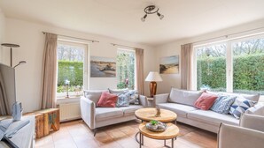 Ein gemütliches Wohnzimmer mit zwei Sofas, einem Couchtisch und einem Fernseher. Große Fenster bieten Blick auf Grünflächen.