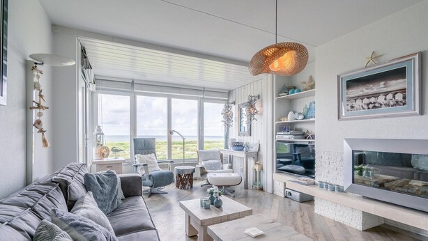 Ein modernes Wohnzimmer mit grauem Sofa, zwei Couchtischen, einem Kamin und einem großen Fenster mit Blick auf eine Rasenfläche.