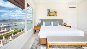 A bedroom with a large window overlooking a marina, a bed with a grey headboard, two bedside tables with lamps, and a bench. - Grand Marina Penthouse (Playa Flamingo)