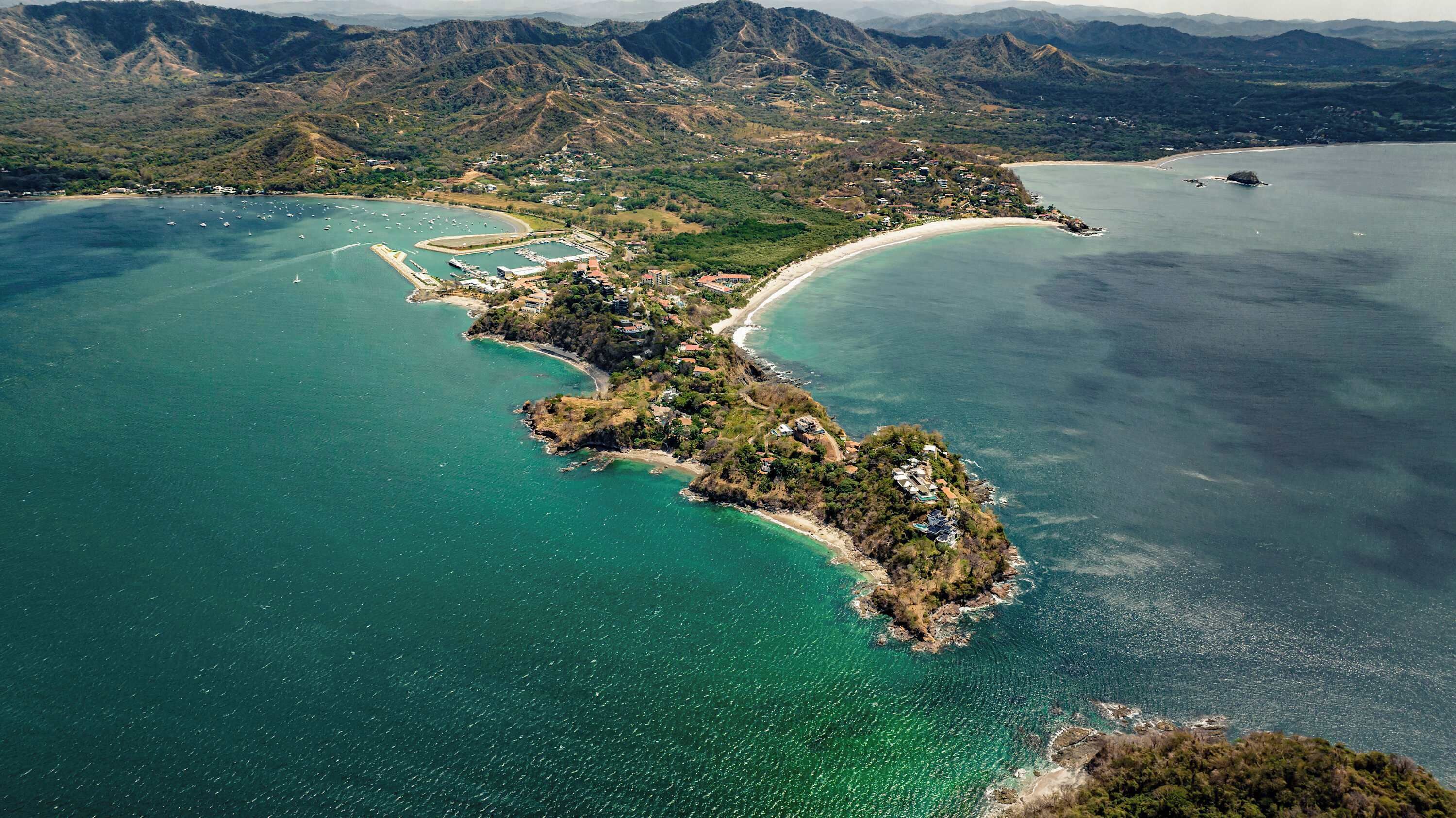 An aerial view of a coastal area with a small peninsula, clear turquoise waters, and a marina with boats.