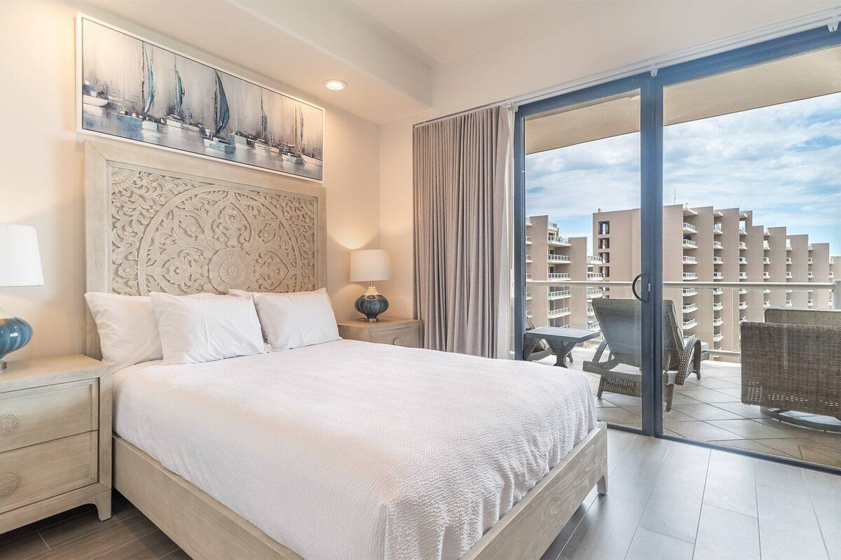 Habitación de hotel con una cama grande, dos mesitas de noche con lámparas, un cabecero decorativo y vistas a la ciudad a través de una puerta corrediza de vidrio.