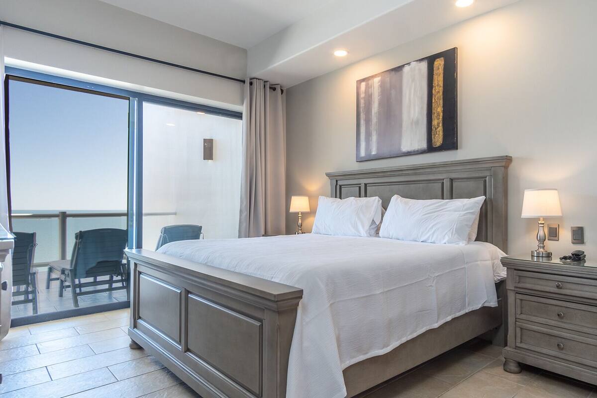 Habitación de hotel con una cama grande, un escritorio y una silla. Tiene un balcón con vistas al mar.