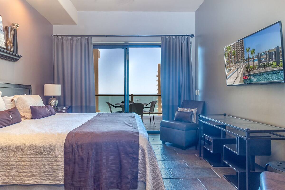Habitación de hotel con una cama grande, un escritorio, una silla y un balcón con vista al mar.