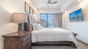 Une chambre à coucher comprenant un lit, une table de chevet, une lampe, un ventilateur de plafond, une fenêtre avec des rideaux et un tableau accroché au mur.