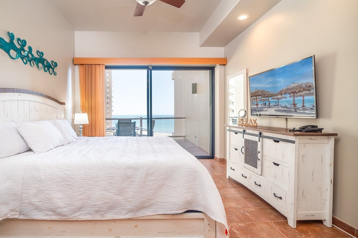 Un dormitorio con cama, mesita de noche, escritorio, televisor y balcón con vista al océano.