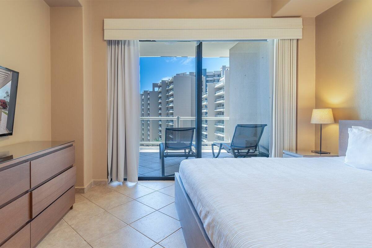 Una habitación de hotel con una cama, un escritorio, una silla, un balcón con vistas a edificios altos y un espejo grande en la pared.