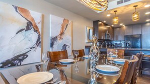 A modern dining area with abstract art on the walls, a glass-top table, and a kitchen in the background.