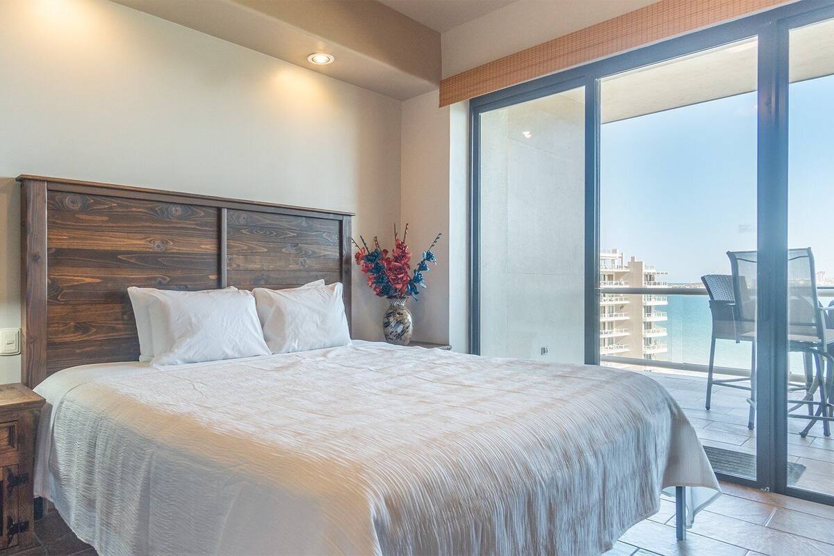 Un dormitorio con cabecera de madera, una mesita de noche con lámpara, una cama grande con ropa blanca, un jarrón con flores y un balcón con vistas a edificios y una masa de agua.