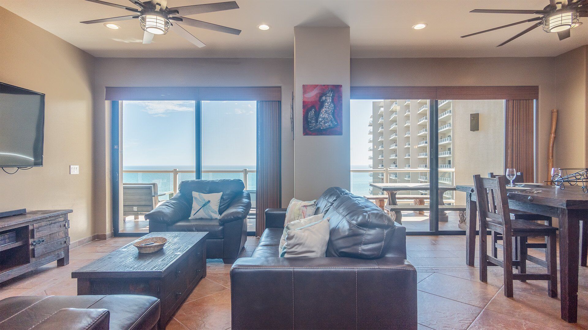 A spacious living area with a leather sofa, a wooden coffee table, and a TV. There is a balcony with a view of a building and the sky.