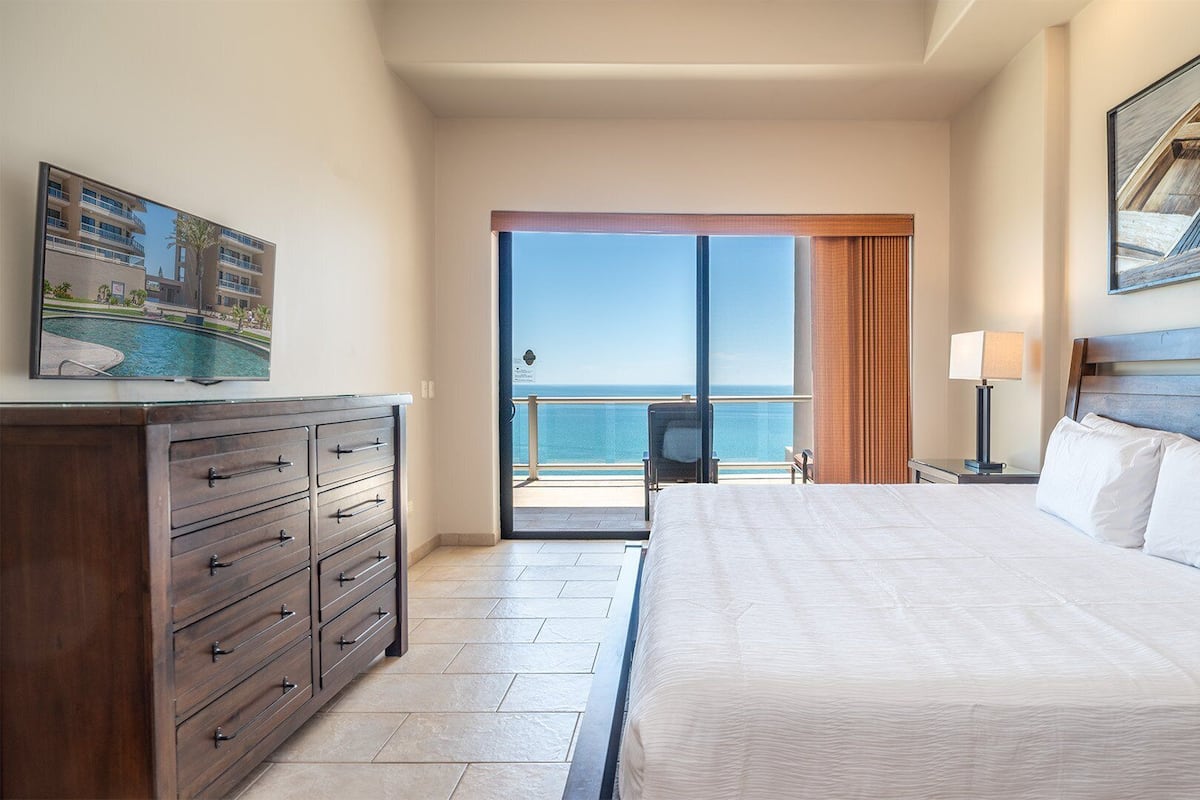 Una habitación de hotel con cama, cómoda, mesita de noche, lámpara y un cuadro de barcos.