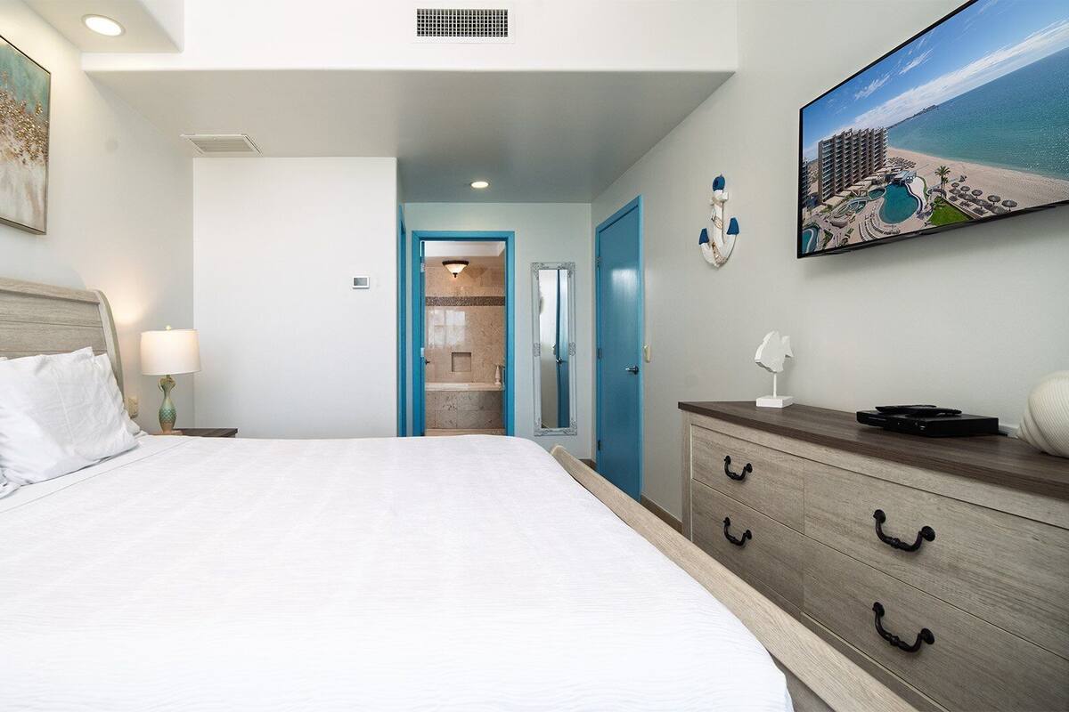 Habitación de hotel con una cama grande, cómoda, un cuadro y vistas a la playa y al hotel desde la ventana.