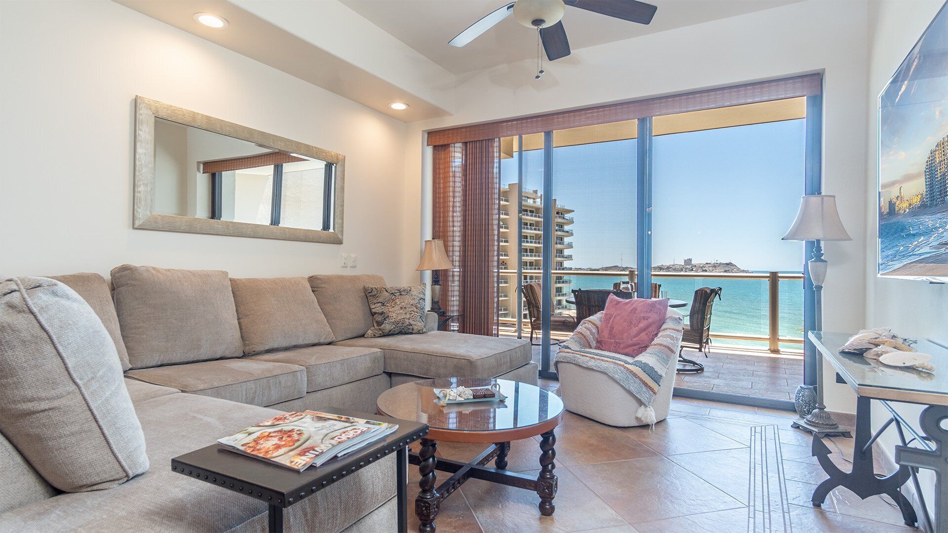 A modern living room with a sectional sofa, a coffee table with magazines, a round table with chairs, and a large window with a view of the sea and buildings.