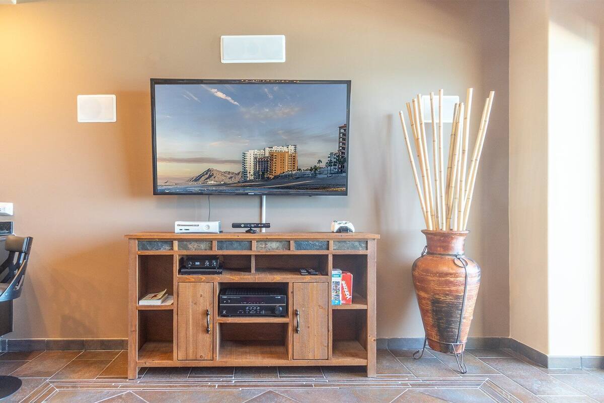 Un vestíbulo de hotel con un televisor montado en la pared, un mueble de madera debajo, una silla y un jarrón grande con ramas.