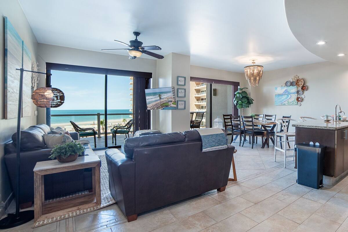 A spacious living area with a view of the beach, a ceiling fan, and a dining area with chairs and a table.