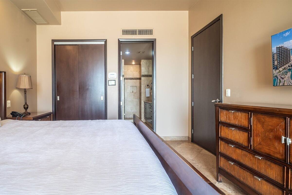 Habitación de hotel con una cama grande, un aparador de madera, una mesita de noche, una lámpara y una puerta que da a un baño.