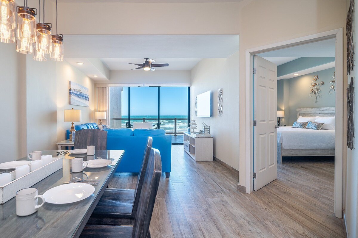 Une chambre d’hôtel moderne dotée d’un coin repas, d’un balcon avec vue sur l’océan et d’une chambre à coucher séparée.