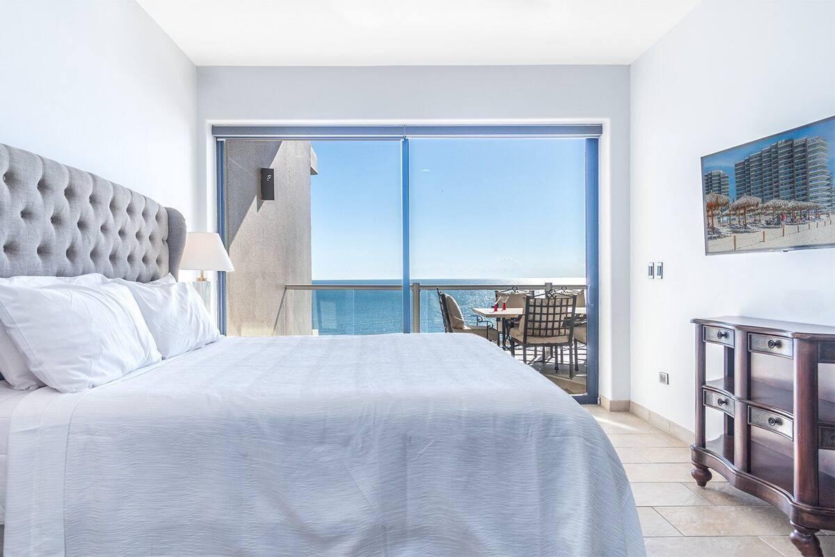 Quarto de hotel com uma cama grande, vista para o mar, varanda com área de estar e um quadro na parede.