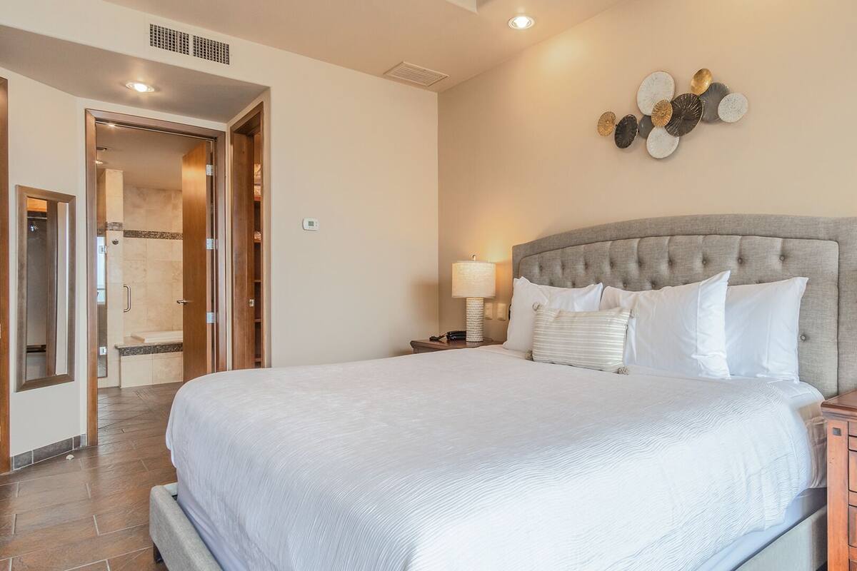 Quarto bem arrumado com uma cama grande, mesas de cabeceira, um relógio de parede e um banheiro visível por uma porta aberta.