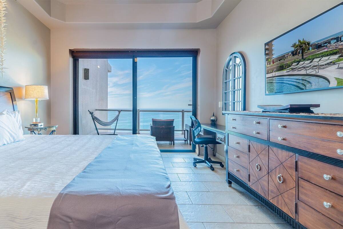 Quarto de hotel com cama, escrivaninha, cadeira, cômoda e vista para o oceano por uma porta deslizante.