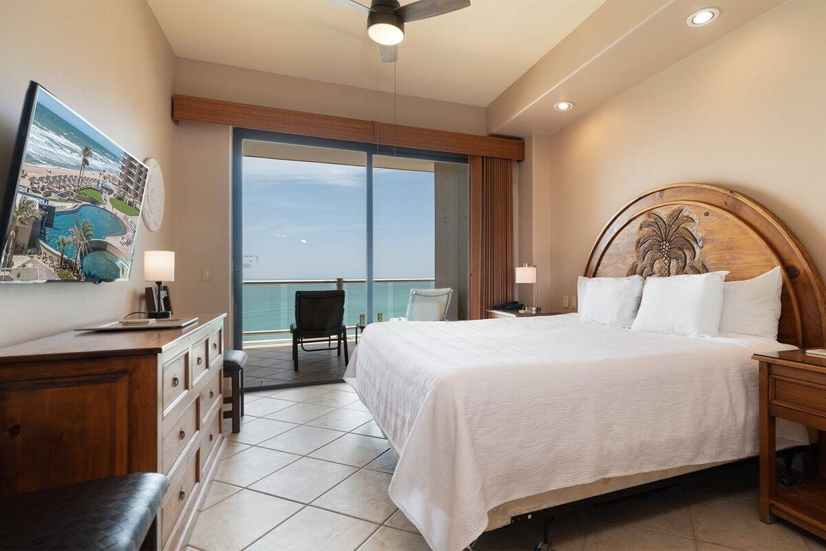 Une chambre d’hôtel comprenant un lit, des tables de chevet, un bureau, une chaise, un balcon avec vue sur l’océan et un tableau au mur.