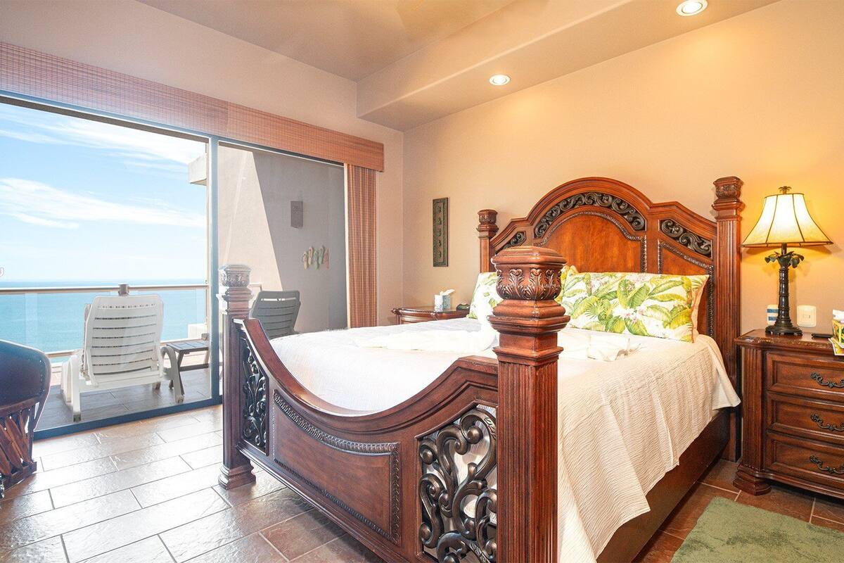 Quarto com cama de madeira, mesa de cabeceira, luminária e vista para o mar através de uma porta de correr.