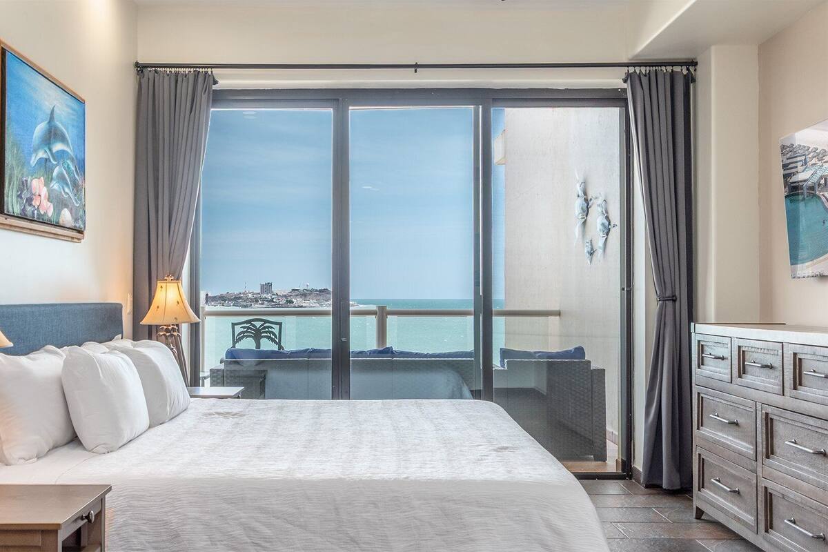 Quarto de hotel com uma cama grande, duas luminárias de cabeceira, vista para o mar e uma varanda com uma cadeira.