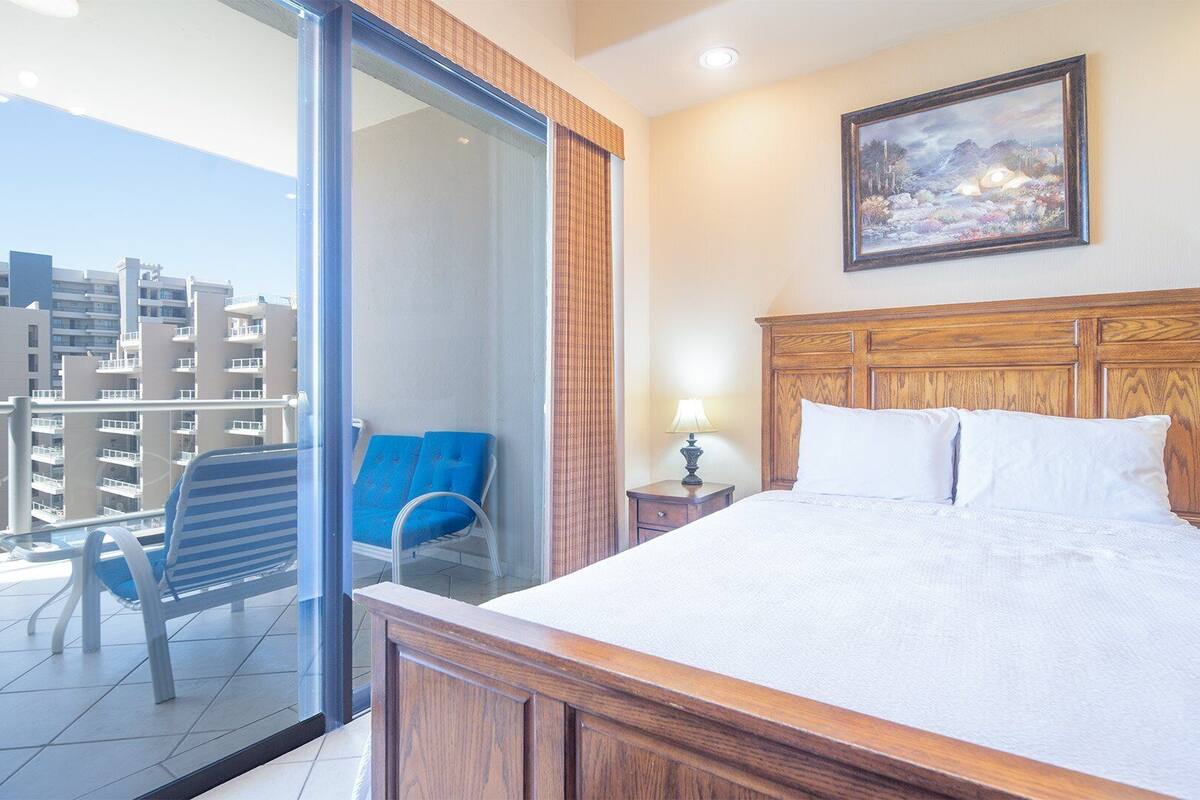 Quarto de hotel com varanda, cama com lençóis brancos, cabeceira de madeira e uma pintura na parede.