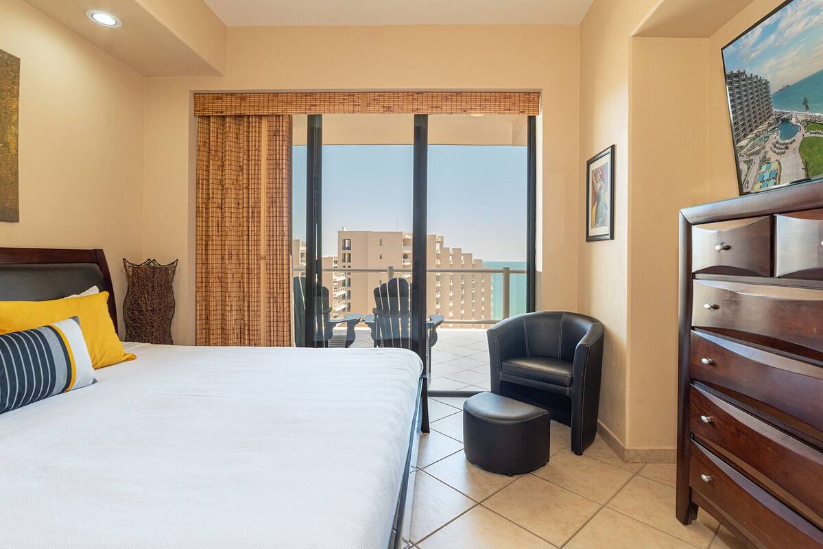 Quarto de hotel com uma cama grande, vista da cidade, varanda com cadeiras e cômoda.