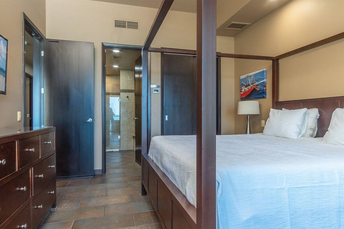 Quarto de hotel com uma cama de dossel, cômoda de madeira, um quadro na parede e porta que dá para o banheiro.