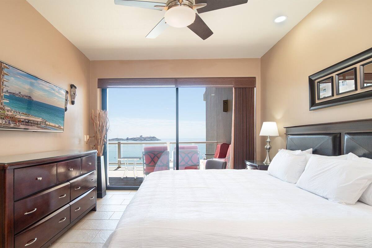 Quarto de hotel com cama grande, ventoinha no teto, pintura de uma paisagem costeira, varanda com cadeiras e vista para o mar.