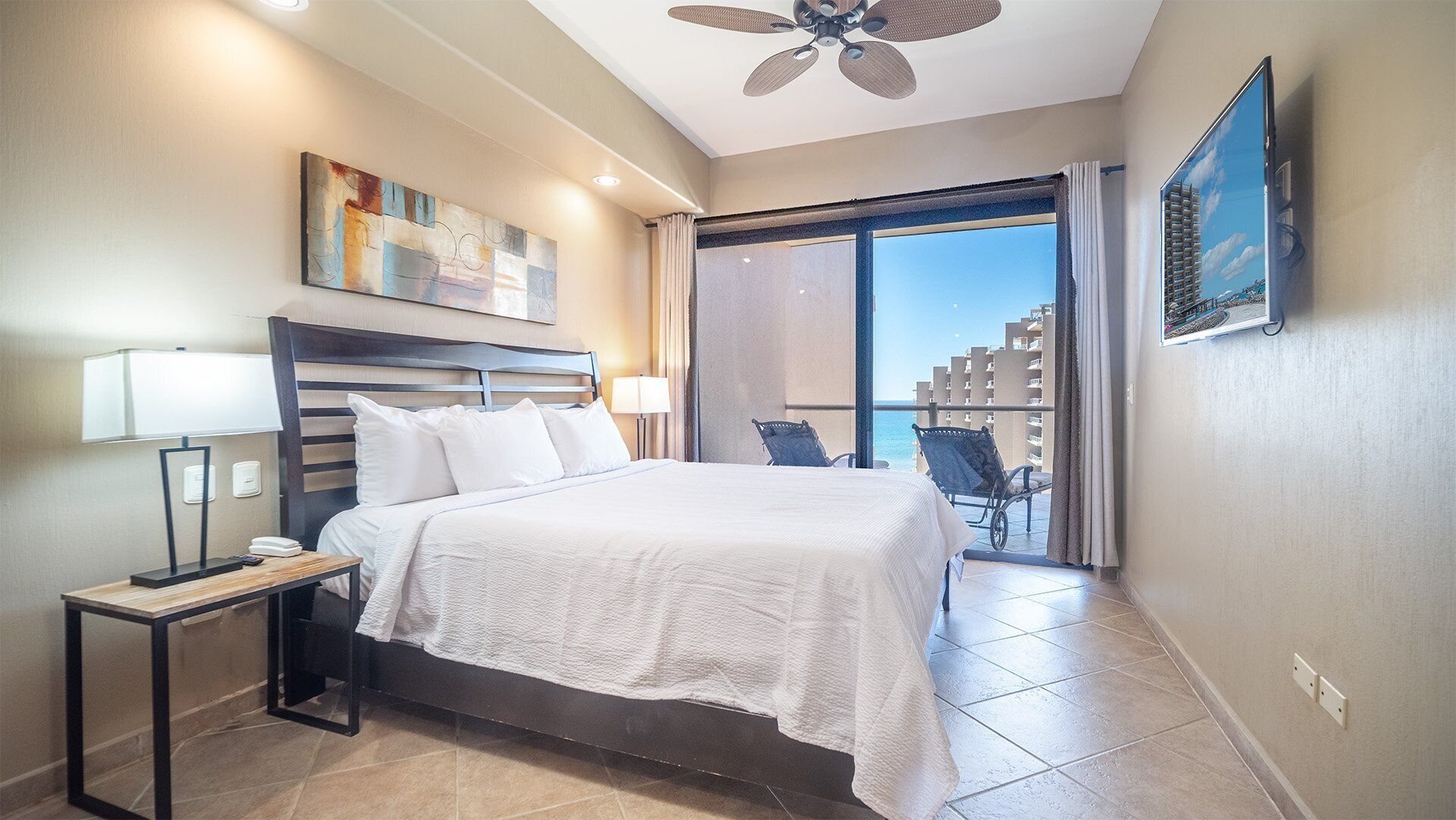 Une chambre d’hôtel comprenant un lit, des tables de chevet, un ventilateur de plafond, une grande fenêtre donnant sur des immeubles et un balcon avec des chaises.