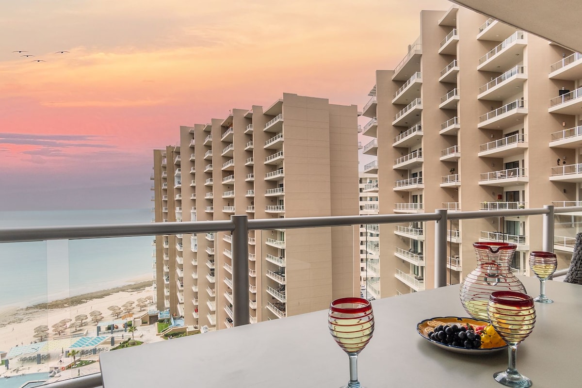 A balcony with a table set for dining, overlooking a beach and high-rise buildings.