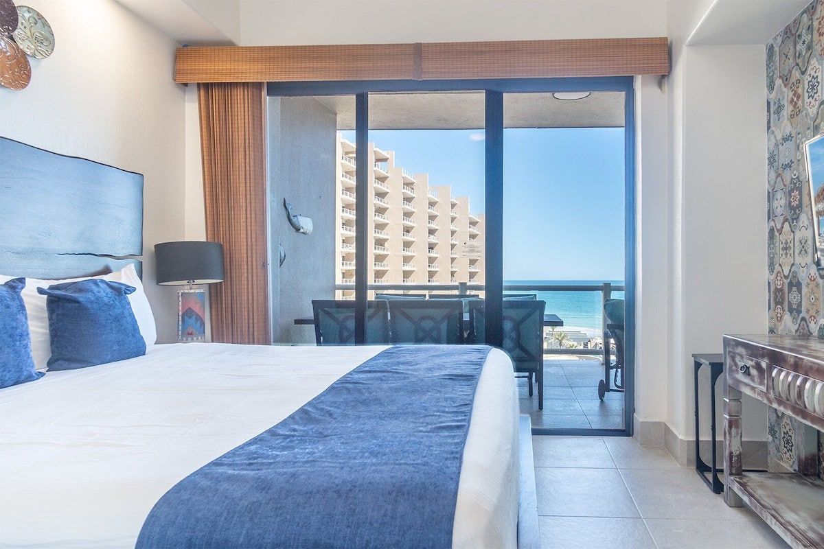 Een hotelkamer met een bed, een bureau, een stoel en uitzicht op de oceaan en een gebouw.
