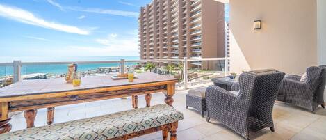 Un balcón con una mesa de madera y sillas de mimbre, con vista a una playa y un edificio de apartamentos.