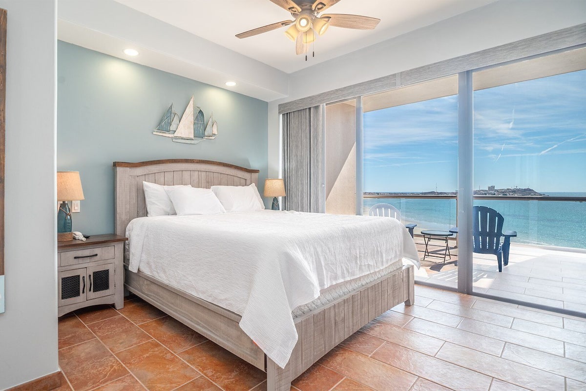 Een slaapkamer met een groot bed, een nachtkastje, een plafondventilator en uitzicht op de zee vanaf een balkon.