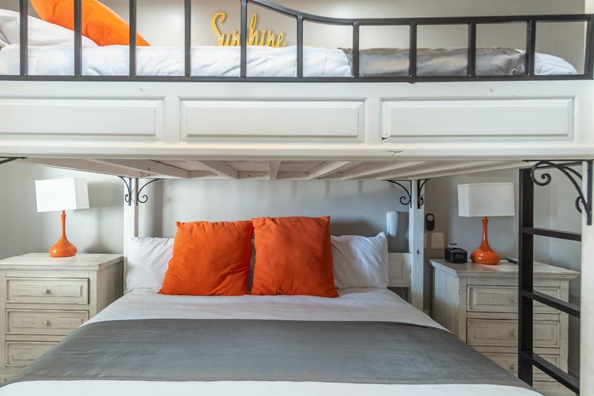 Een tweepersoonsbed met een grijze spreidek en twee oranje kussens, met aan weerszijden witte nachtkastjes met lampen, in een kamer met een zwart-wit kleurenschema.