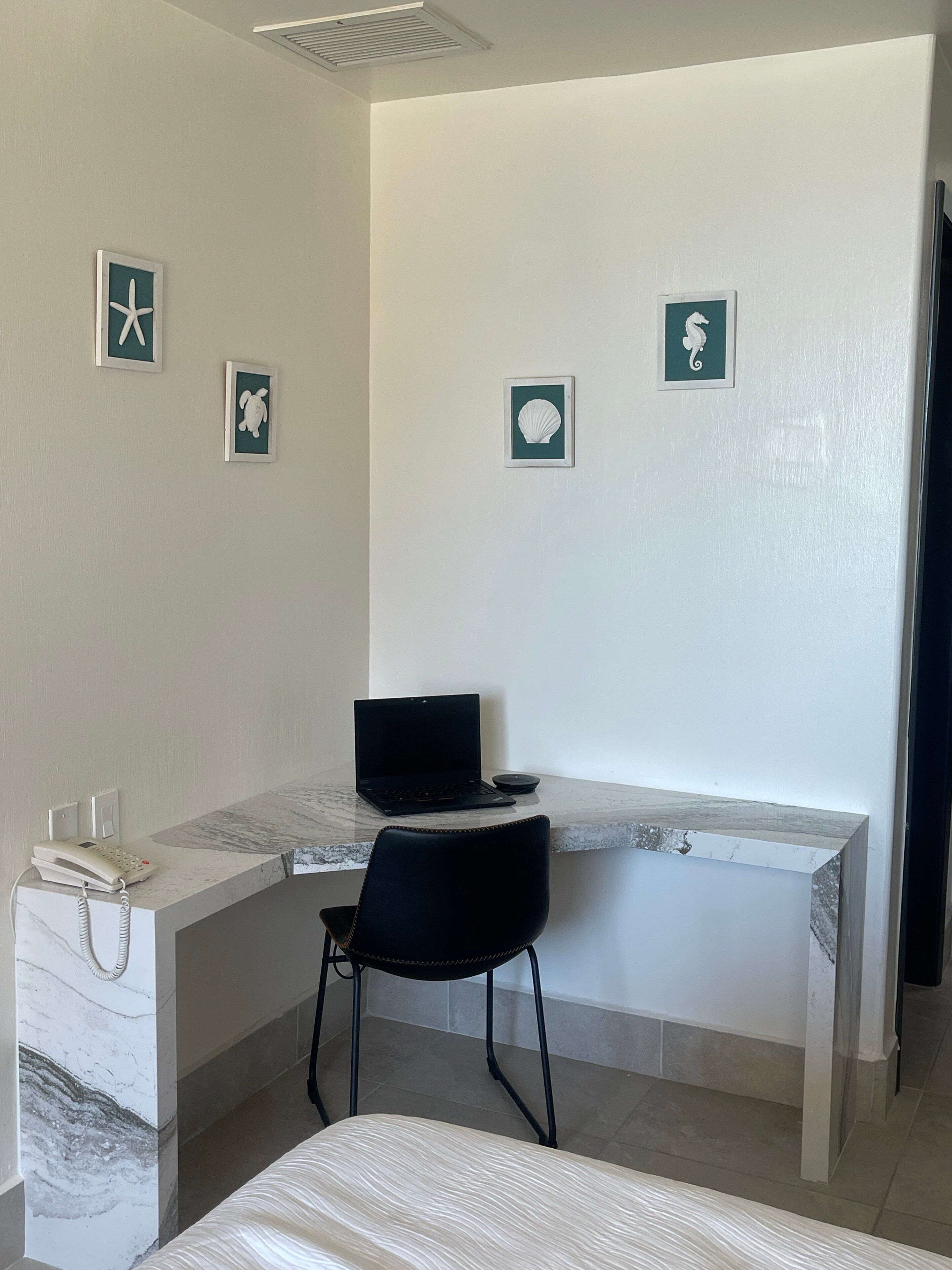 Une chambre d’hôtel avec un lit, un bureau avec un ordinateur portable, une chaise et un téléphone. Des tableaux représentant une tortue, une coquille et un hippocampe sont accrochés aux murs.