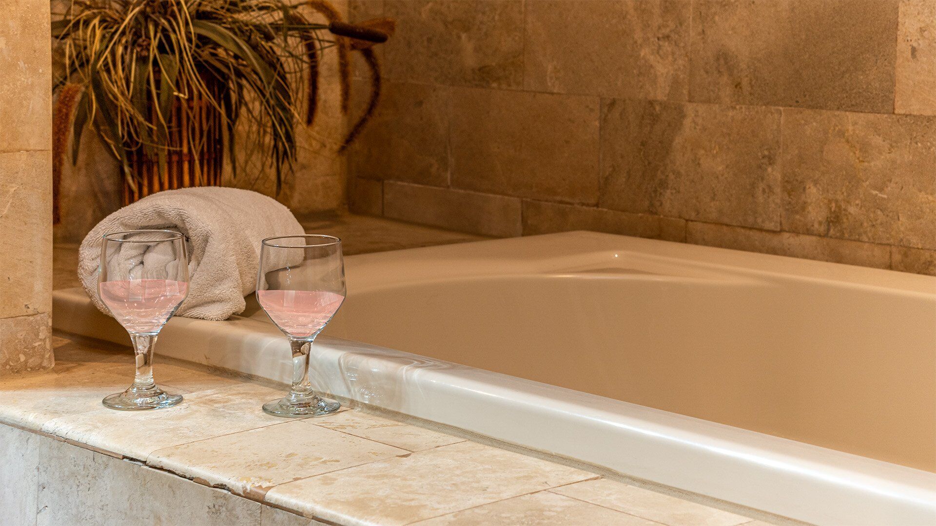 A bathtub with two glasses of pink liquid, a towel, and a plant in a bathroom setting.