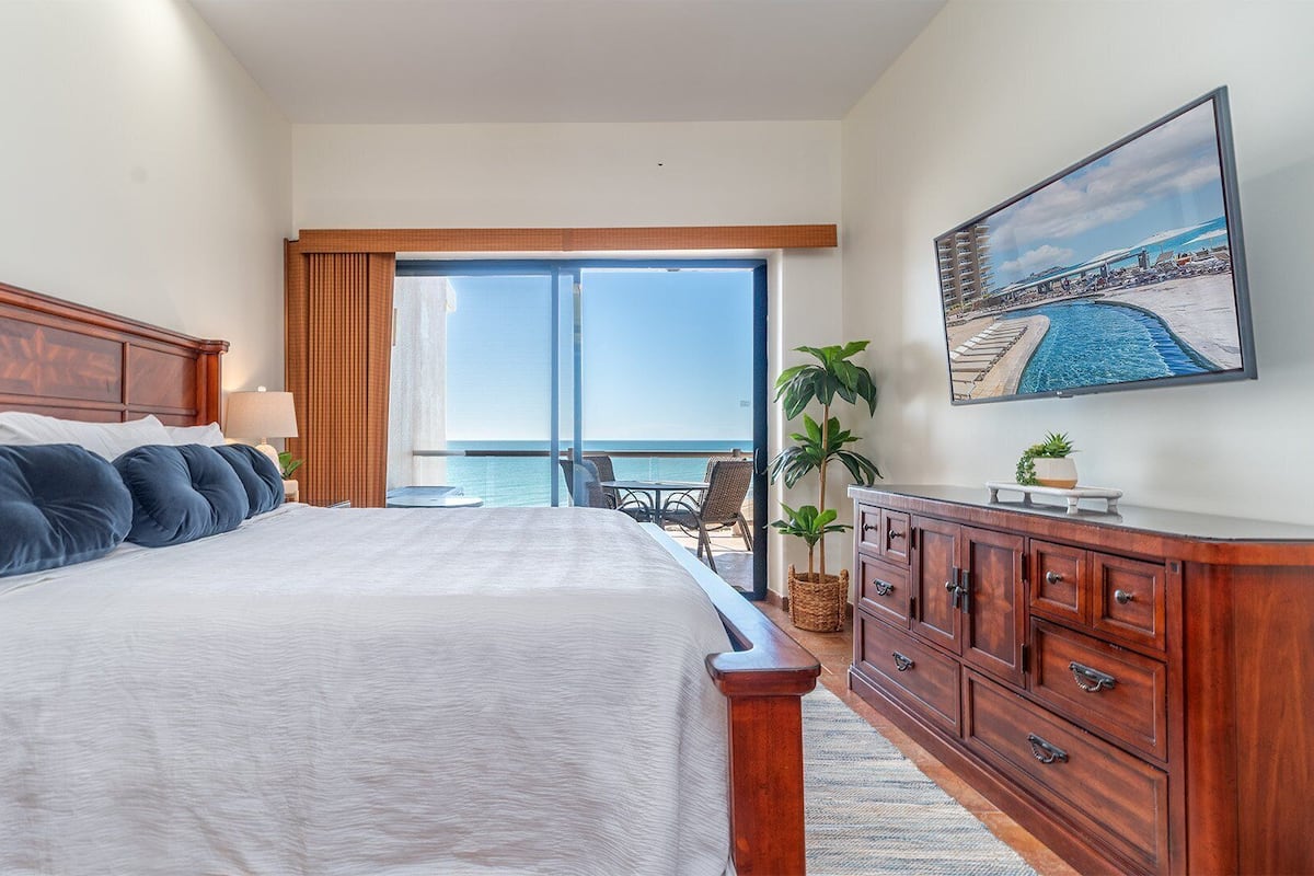 Een slaapkamer met een groot bed, een houten hoofdeinde, een nachtkastje, een dressoir, een televisie en uitzicht op de oceaan door een schuifdeur.
