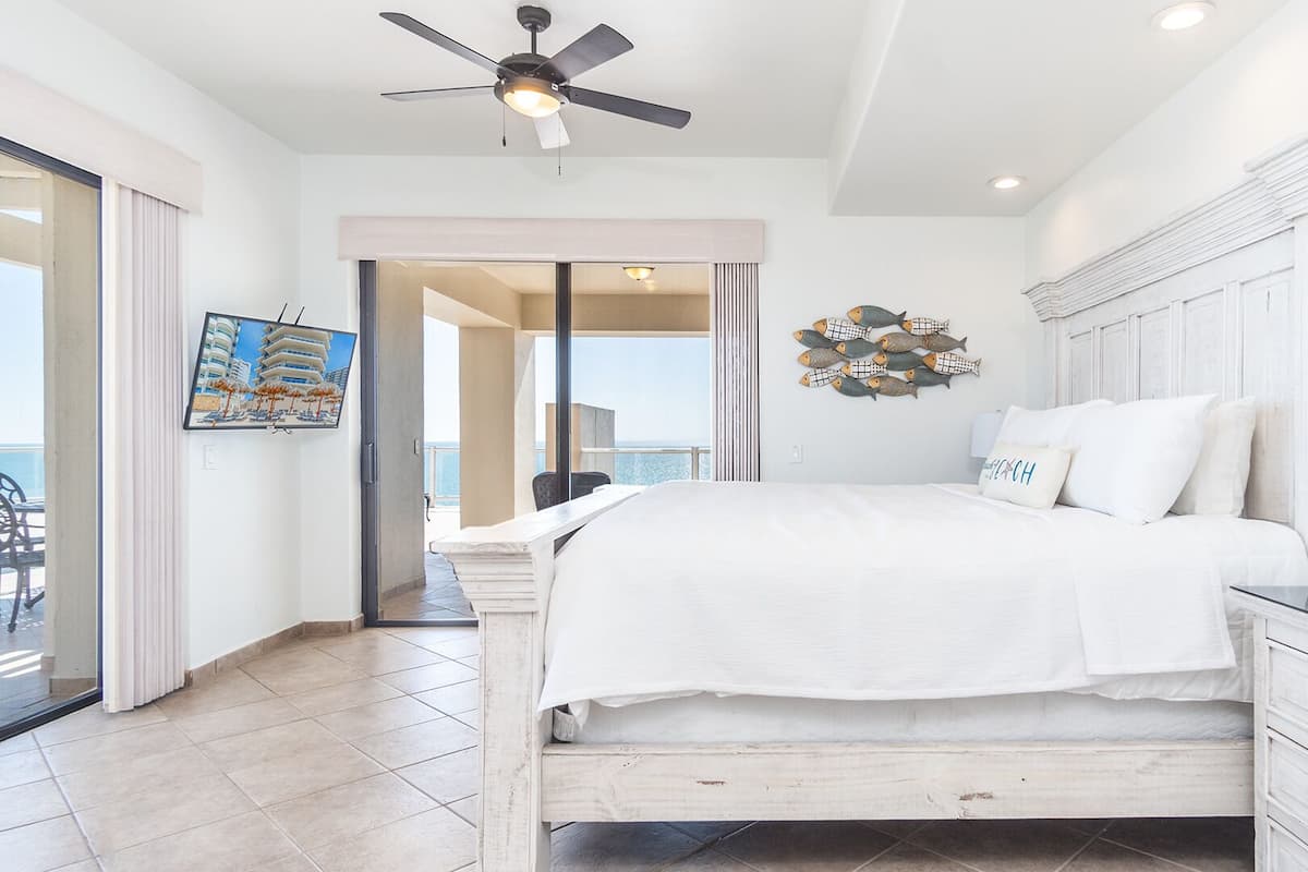 A bedroom with a bed, a ceiling fan, a wall-mounted fish decoration, and a view of the sea through a sliding door.