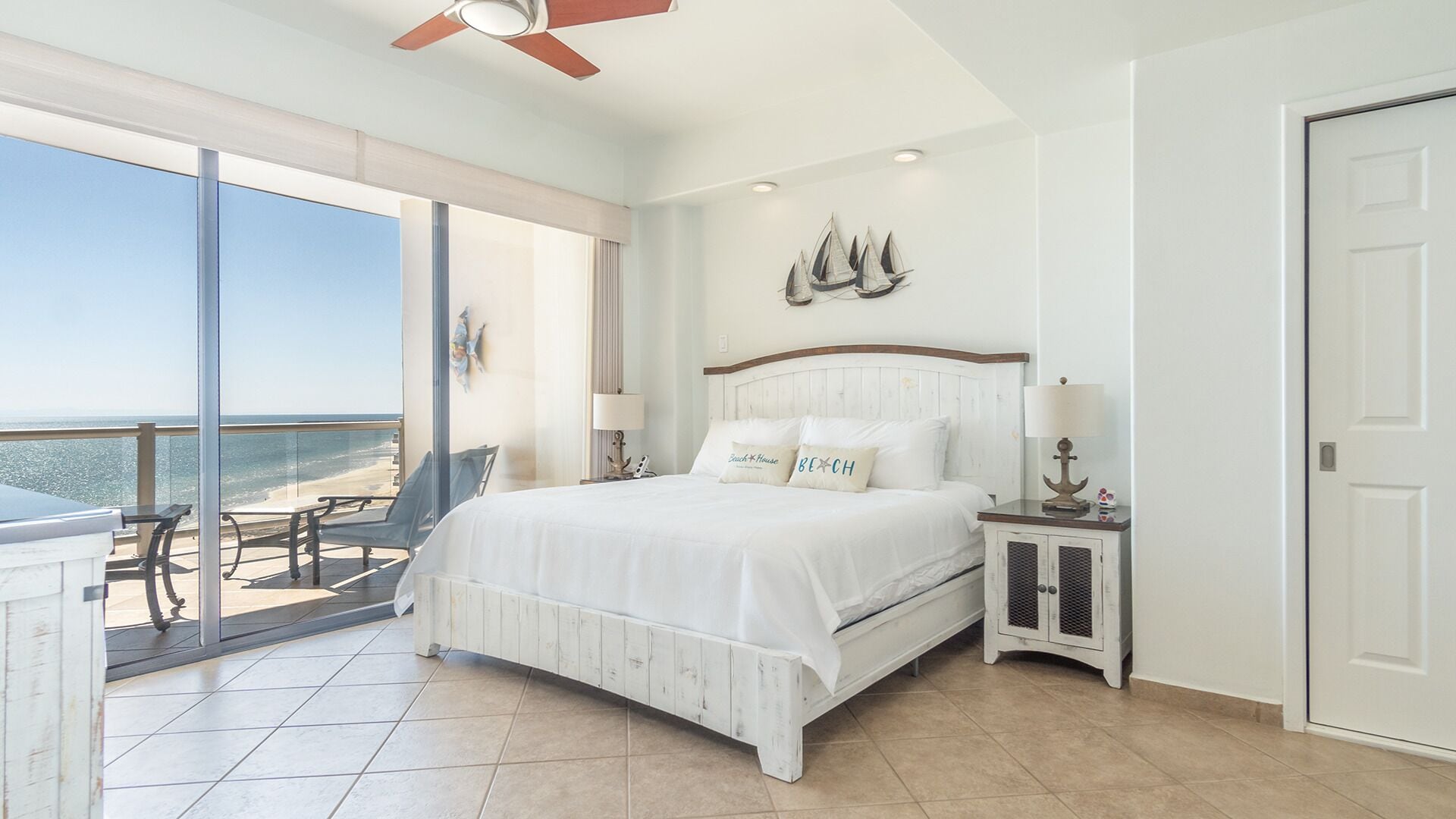 Quarto com uma cama grande, uma mesa de cabeceira, uma luminária e vista para a praia através de uma porta de vidro deslizante.