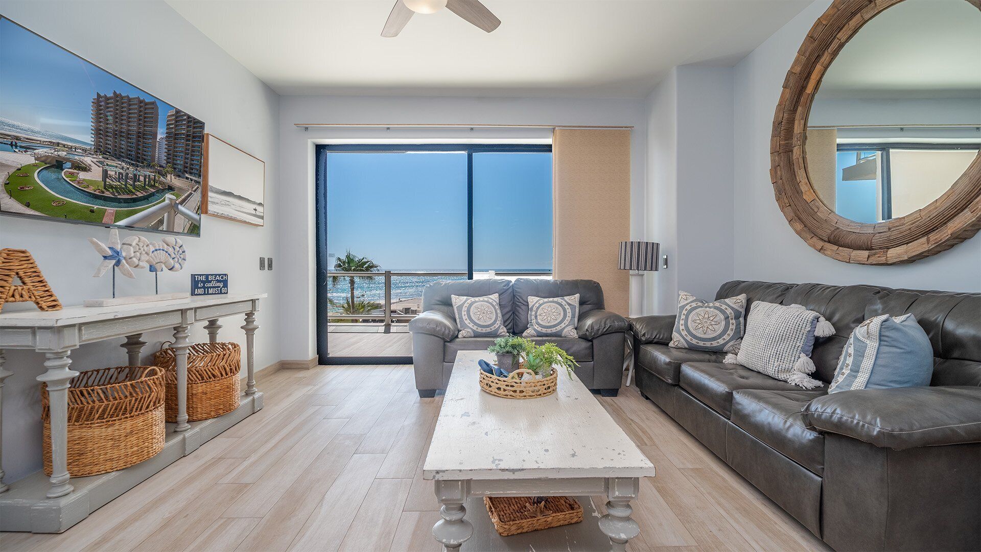 A spacious living room with a large window overlooking a beach, a comfortable gray sofa, and a white coffee table with decorative items.