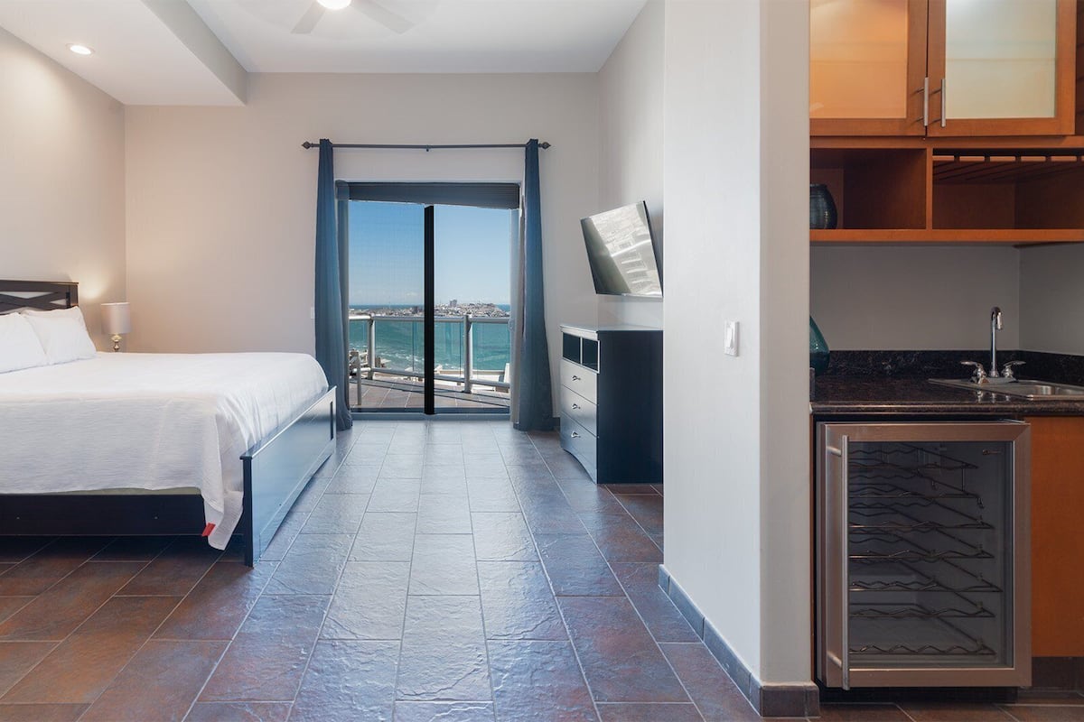 Habitación de hotel moderna con cama, televisor, una kitchenette pequeña y un balcón con vista al mar.