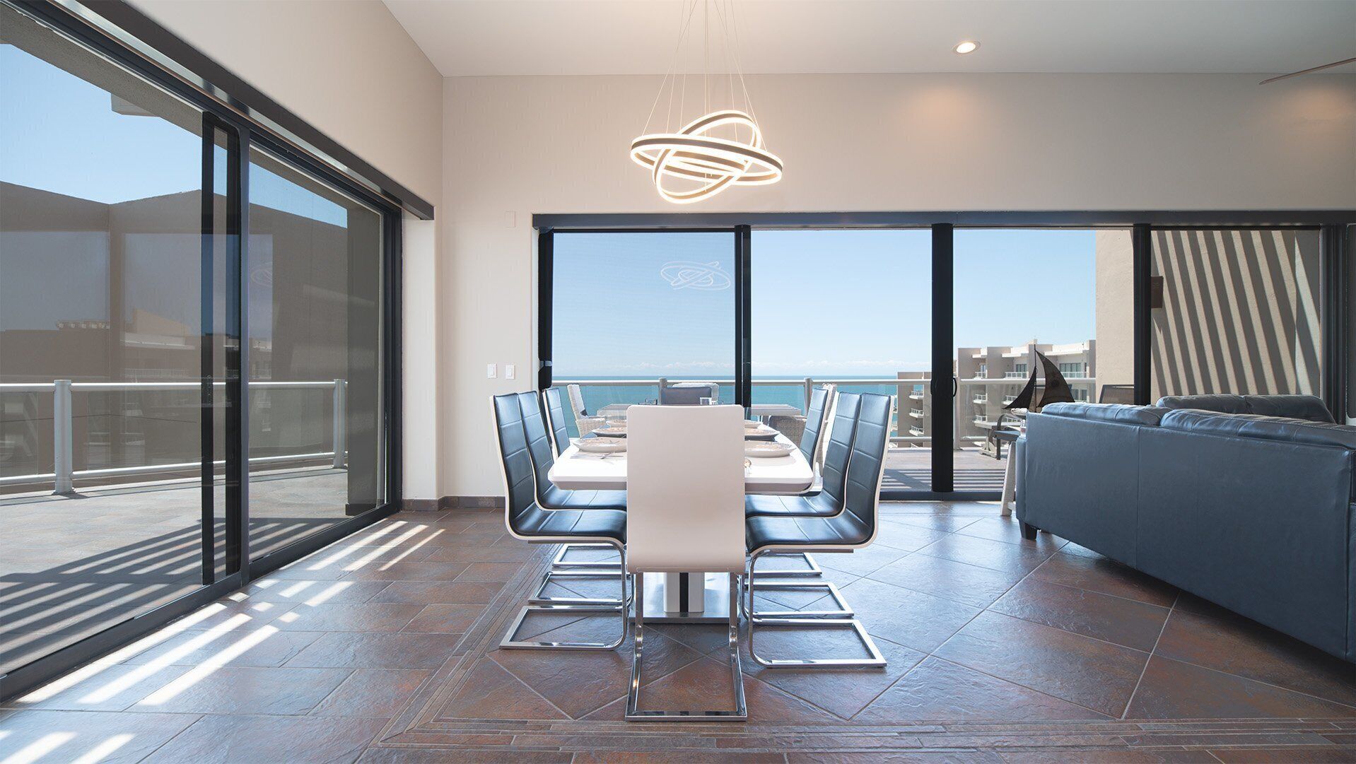 A modern dining area with a table and chairs, a sofa, and a large window offering a view of the sea.