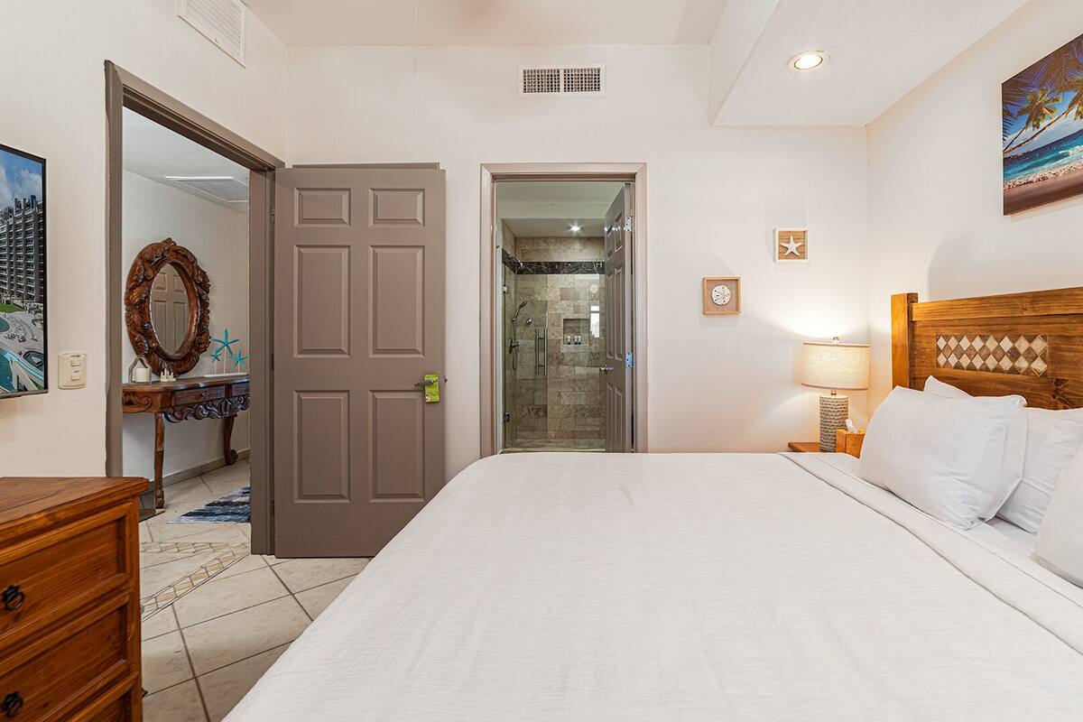 Une chambre d’hôtel avec un lit, une commode en bois, une salle de bain avec douche et un tableau représentant une scène de plage.