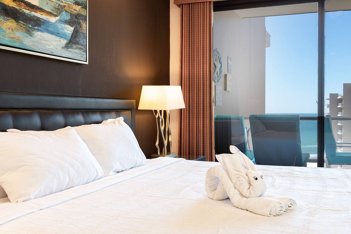 Een hotelkamer met een groot bed, uitzicht op zee en een balkon met stoelen.