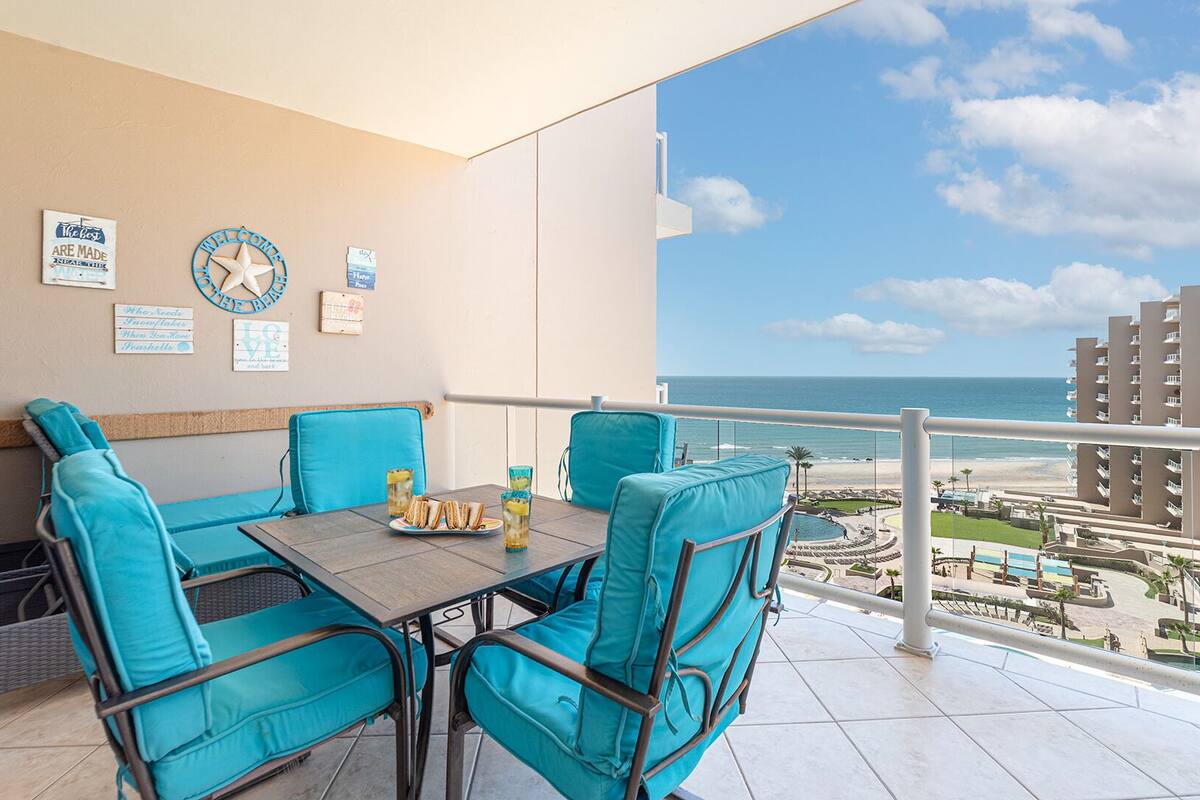 Un balcon avec des chaises turquoise et une table, d’où l’on a vue sur une plage et les bâtiments d’un hôtel.