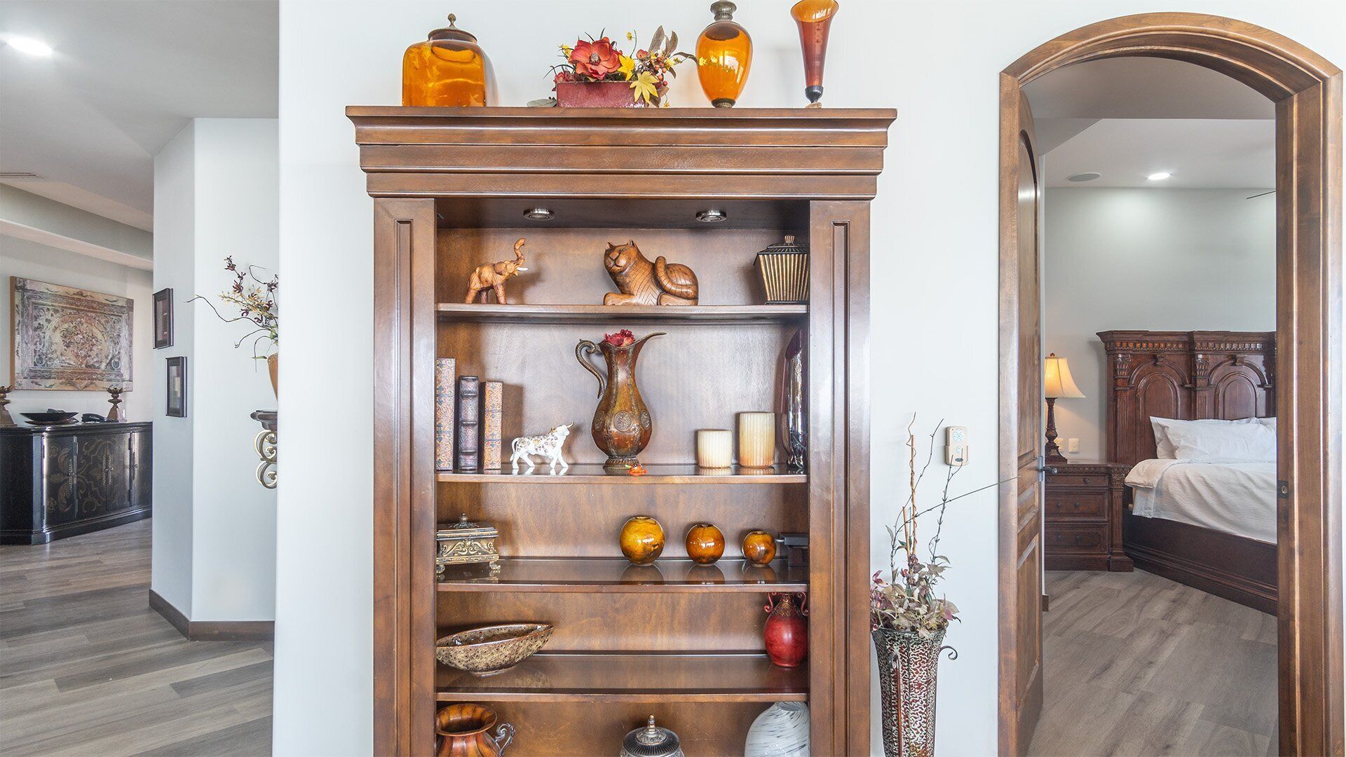 Un mueble de madera con repisas que contienen objetos decorativos, un espejo y una cama en la habitación contigua.